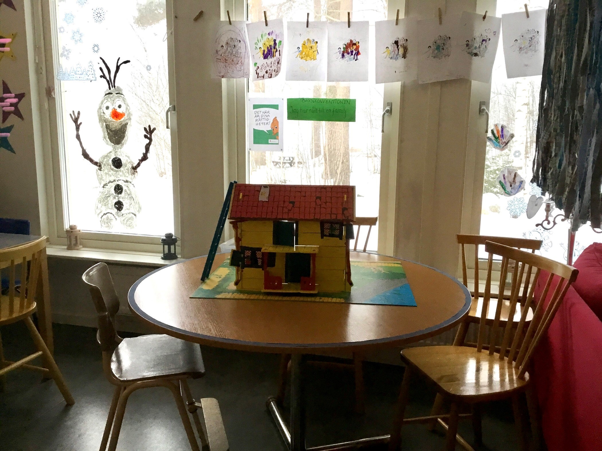 Glad snögubbe målad på fönstret. Runt bord med stolar, på bordet står ett gult litet hus. Teckningar hänger i klädnypor på en lina.