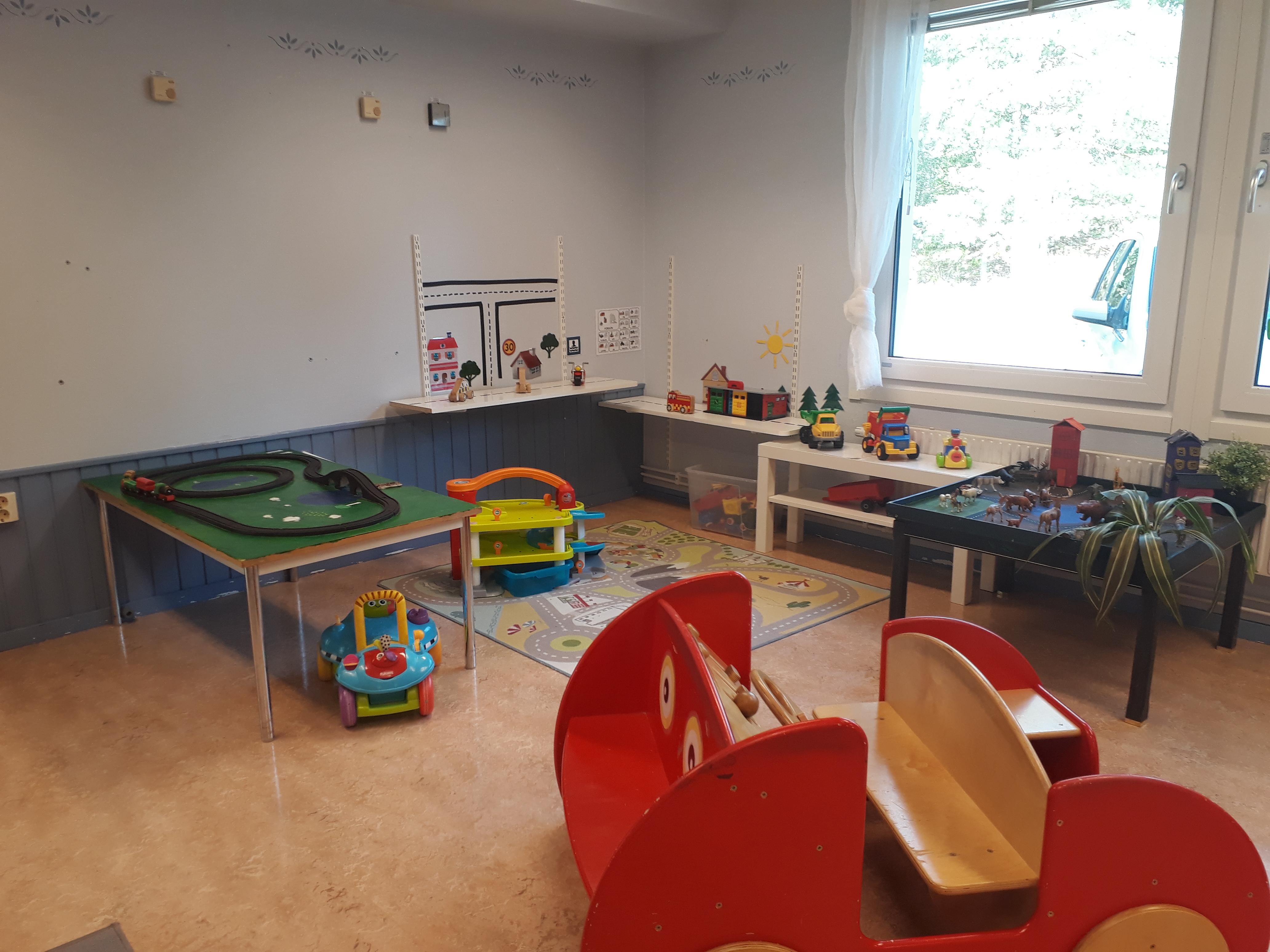 Ett bord med en tågbana. På golvet ligger en bilmatta och på den står det ett leksaksgarage. En röd leksaksbil som barnen kan sitta i.
