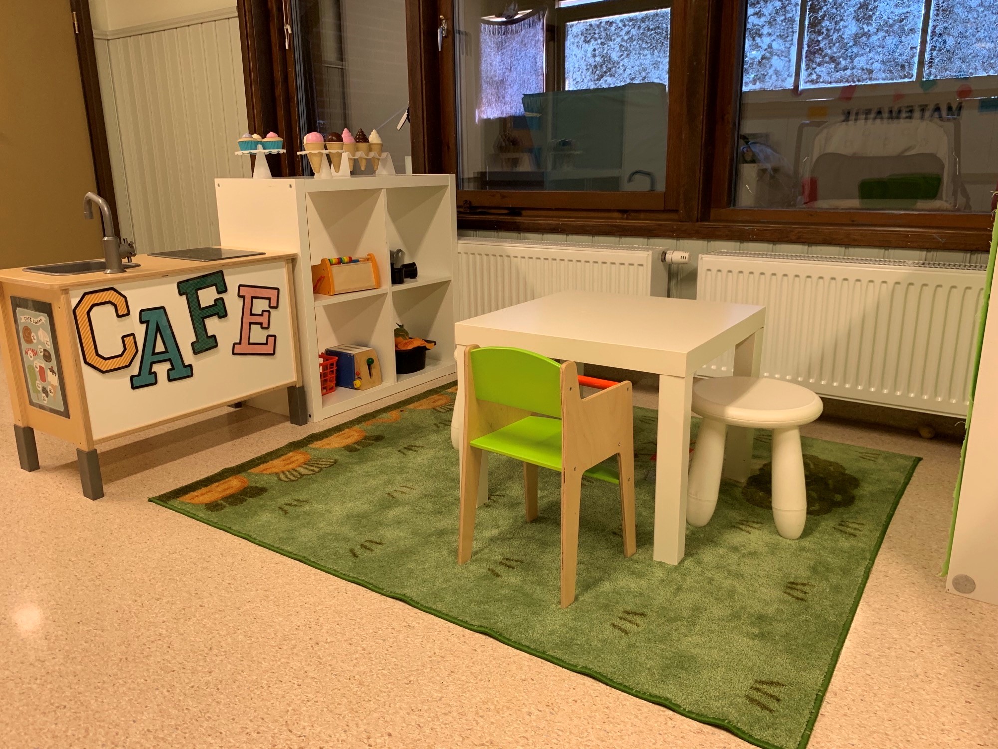 Ett vitt litet fyrkantigt bord med 2 vita pallar och en stol, står på en grön matta. Ett leksakskök finns till vänster med diskho. Bakverk i plast står på en vit hylla.