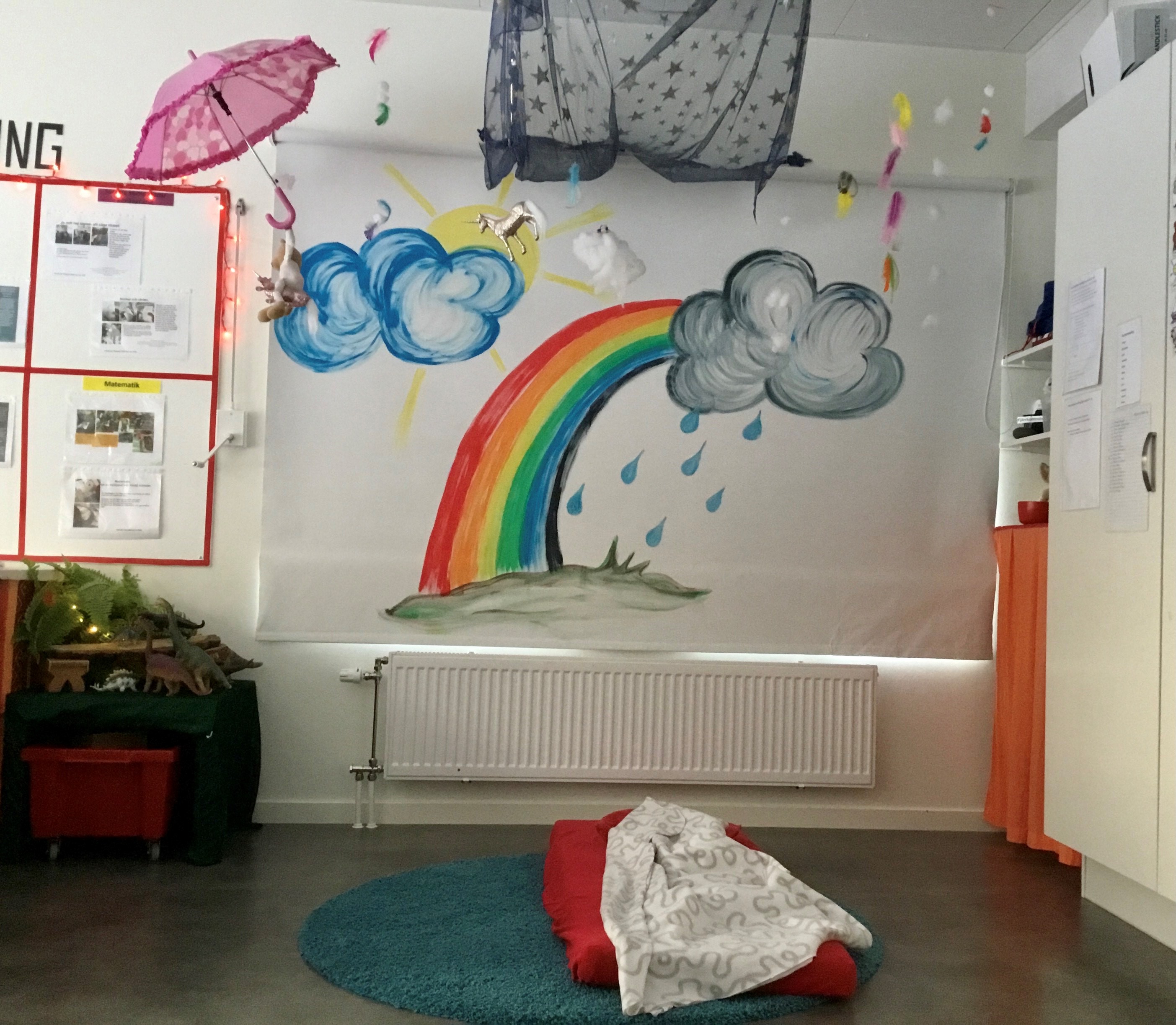 En regnbåge är målad på väggen. Ett grått regnmoln i toppen på regnbågen. Till väsnter om regnbågen är det ett blått moln med en sol bakom. Ett rosa paraply hänger i taket