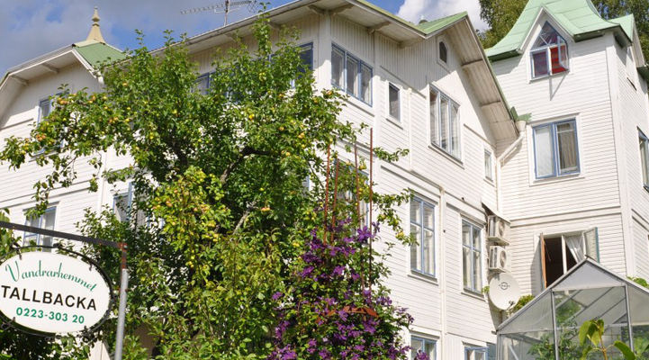 Vandrarhemmet Tallbacka i Ängelsberg. En vit villa med grönt plåttak. Grönska med träd omringar villan