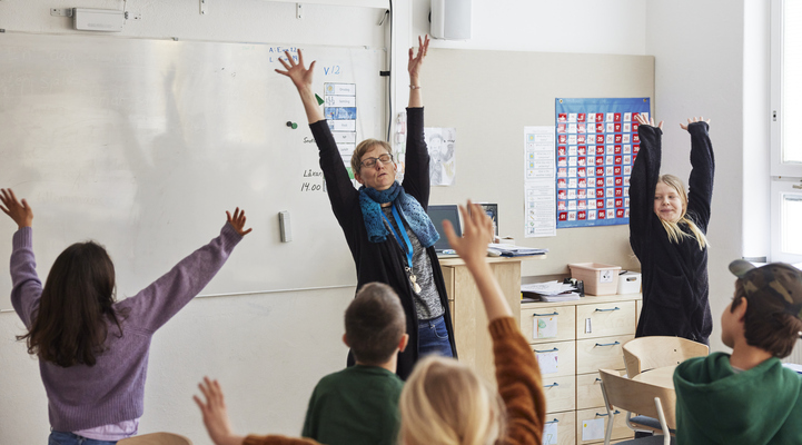 Äldre lärare står längst fram vid whiteboardtavlan och håller sträcker upp armarna inför klassen. Eleverna gör lika som läraren.
