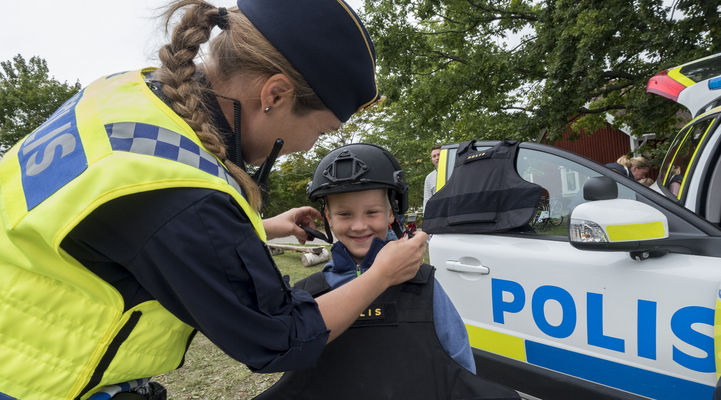 En kvinnlig polis hjälper en pojke att knäppa en polishjälm när han testar polisens utrustning. Till höger i bild så synd en del av en polisbil.