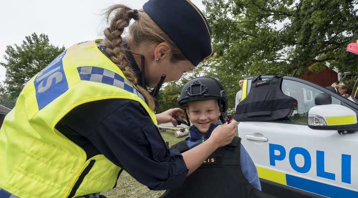 En kvinnlig polis hjälper en pojke att knäppa en polishjälm. Pojken testar polisens utrustning. Till höger i bild syns en del av en polisbil.