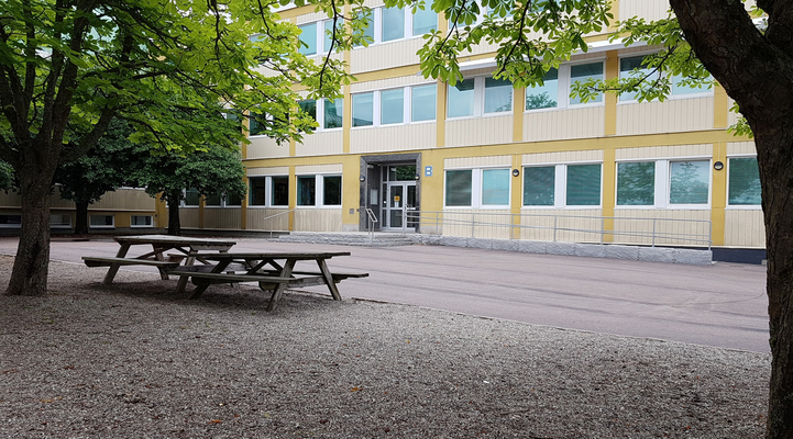 Del av Brinellskolans byggnad. Mellan två träd skymtas entrén till en gul byggnad. I förgrunden står två picknickbord under ett av träden.