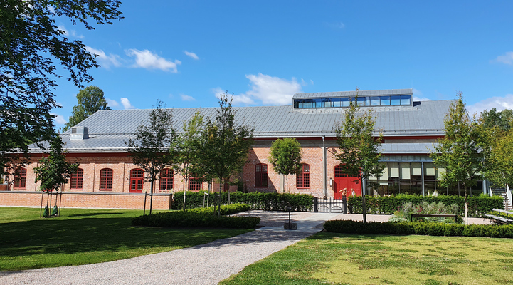 Konferensanläggning. En tegelbyggnad med röda tak och silverfärgat plåttak. I förgrunden syns gräsmatta, en grusväg och nyplanterade träd.