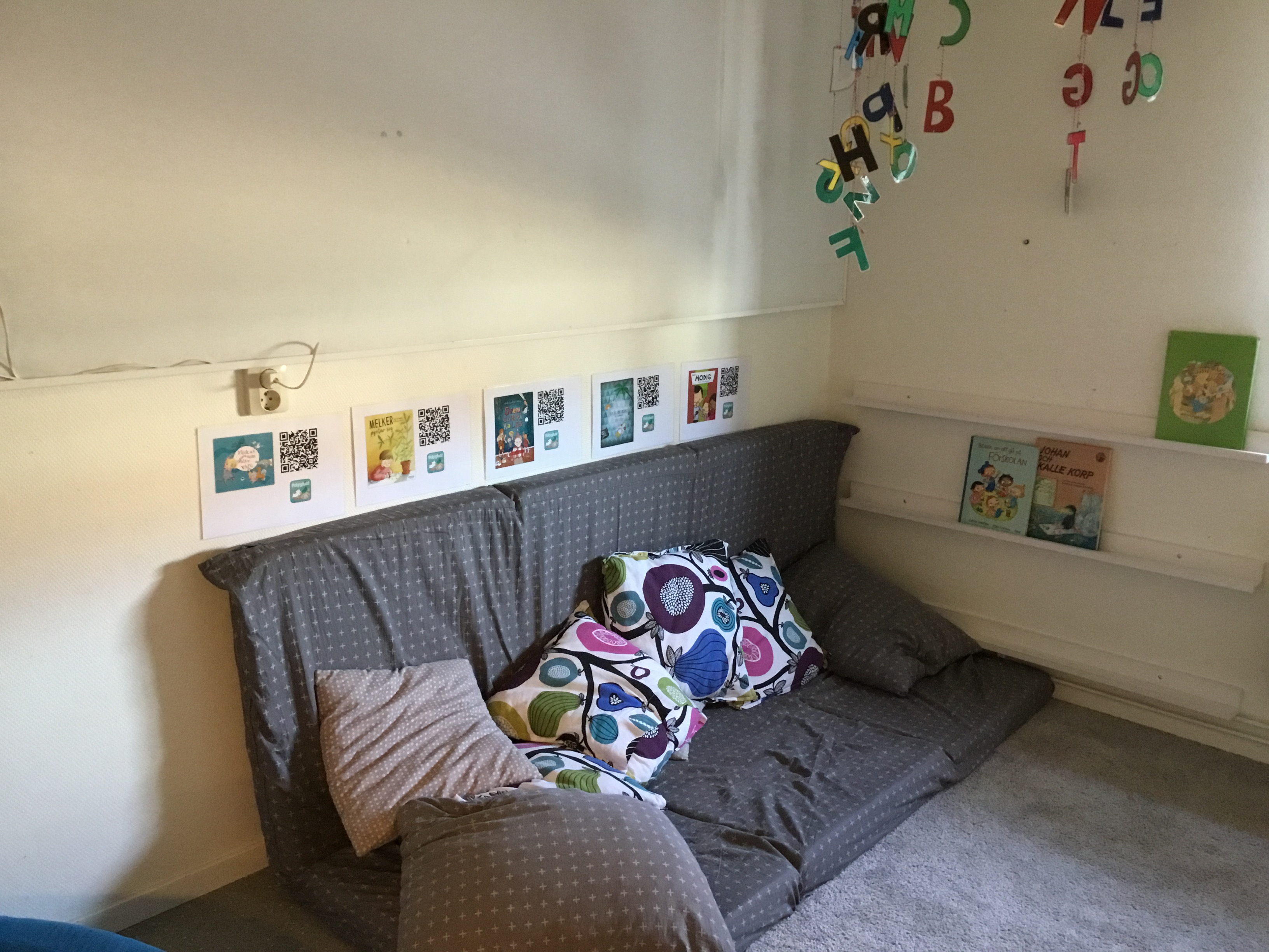 En grå låg soffa med kuddra, Bokstäver sitter på väggarna coh teckningar hänger över soffan.